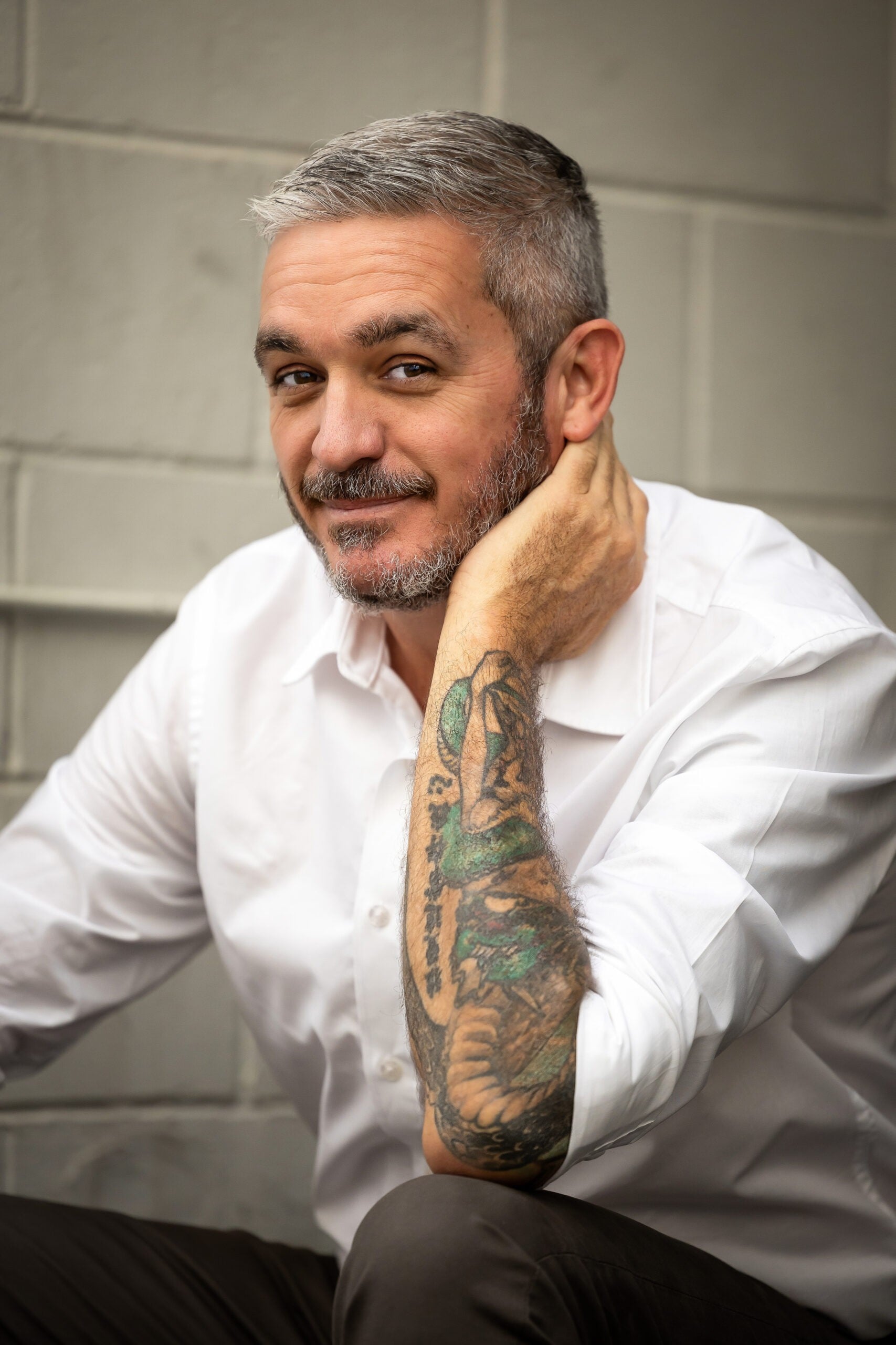 A man with short grey hair and a beard, wearing a white shirt, sits in front of a light brick wall. Smiling warmly and resting his head on his hand, he reveals a colourful tattoo—exuding the relaxed vibe of a tantric massage Australia expert.